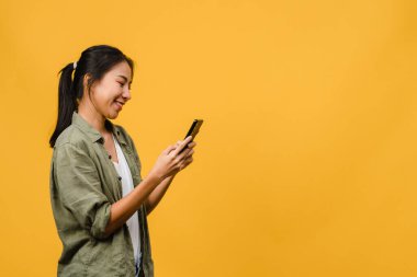 Genç Asyalı bayan telefonu olumlu bir ifadeyle kullanıyor, geniş bir şekilde gülümsüyor, gündelik giysiler içinde mutluluğu hissediyor ve sarı arka planda tek başına duruyor. Mutlu, mutlu, mutlu kadın başarıya sevinir..