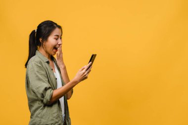 Genç Asyalı bayan telefonu olumlu bir ifadeyle kullanıyor, geniş bir şekilde gülümsüyor, gündelik giysiler içinde mutluluğu hissediyor ve sarı arka planda tek başına duruyor. Mutlu, mutlu, mutlu kadın başarıya sevinir..