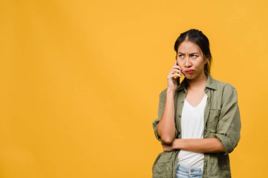 Genç Asyalı bayan telefonda olumsuz bir ifadeyle konuşuyor, çığlık atıyor, duygusal olarak ağlıyor, gündelik giysiler içinde öfkeli ve boş fotokopi çekilen sarı arka planda yalnız duruyor. Yüz ifadesi kavramı.