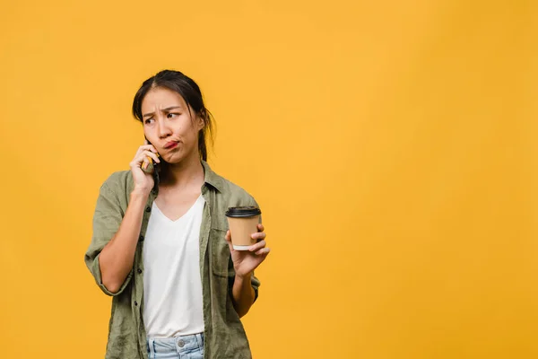 Young Asia lady talk by phone and hold coffee cup with negative expression, excited screaming, cry emotional angry in casual cloth and stand isolated on yellow background. Facial expression concept.