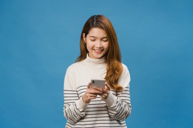 Genç Asyalı bayan telefonu olumlu bir ifadeyle kullanıyor, geniş bir şekilde gülümsüyor, gündelik giysiler içinde mutluluğu hissediyor ve mavi arka planda tek başına duruyor. Mutlu, mutlu, mutlu kadın başarıya sevinir..