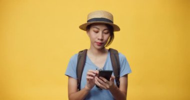 Mutlu, genç, güzel Asyalı bir kadın şapka takıyor ve sırt çantalı akıllı bir telefon kullanıyor. Seyahat oteli ve bilet defterinin keyfini çıkarın. Gülümseyen kız heyecanla sarı arka plandaki boş reklama bakıyor..