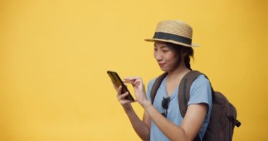 Mutlu, genç, güzel Asyalı bir kadın şapka takıyor ve sırt çantalı akıllı bir telefon kullanıyor. Seyahat oteli ve bilet defterinin keyfini çıkarın. Gülümseyen kız heyecanla sarı arka plandaki boş reklama bakıyor..
