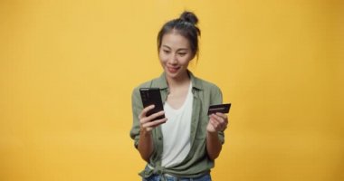 Güzel Asya 'lı yalnız genç bayan, rahat gömlekli, kameraya bakın akıllı telefon ve kredi kartı alışverişinin keyfini çıkarın ve sarı hardal arkaplan üzerindeki başarı ödemesiyle heyecanlanın..