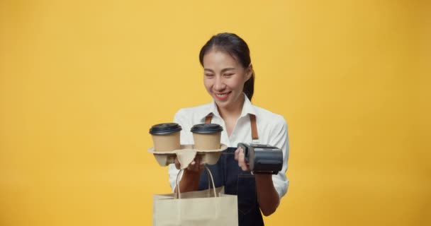 黄色の背景に隔離された顧客にコーヒーや紅茶やクレジットカードリーダーマシンを与えるカメラを見てフレンドリーなエプロンを身に着けている幸せな笑顔アジアのウエートレス ビジネステイクアウトのコンセプト — ストック動画