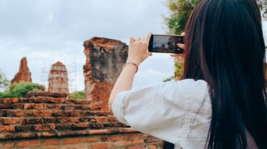 Gezgin Asyalı kadın fotoğraf çekmek için akıllı telefon kullanıyor. Tatil gezisini Ayutthaya, Tayland 'da geçiriyor. Japon bayan turist, geleneksel kentteki inanılmaz şehir simgesinde yolculuğunun tadını çıkarıyor..