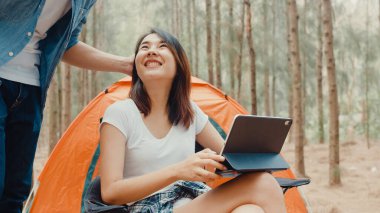 Genç Asyalı kampçılar ormanda çadırın yanında tablet kullanıyorlar. Erkek ve kadın gezgin bir yaz günü kamp alanında dinlenip sohbet ediyorlar. Açık hava aktivitesi, macera seyahati veya tatil tatili.