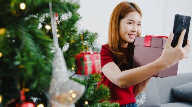Genç Asyalı bir kadın akıllı bir telefonla X 'Mas hediye kutusuyla konuşan bir çiftle konuşuyor. Evdeki oturma odasında süslerle süslenmiş bir Noel ağacı. Noel ve Yeni Yıl bayramı festivali.