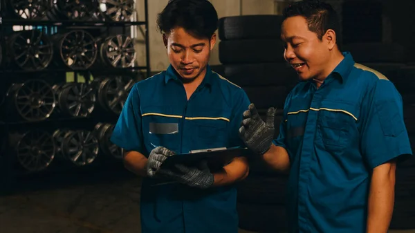 Two professional car mechanic using paperwork makes the oil and engine check to the car on lifted automobile at repair service station at night. Skillful Asian guy in uniform fixing car. Car service.
