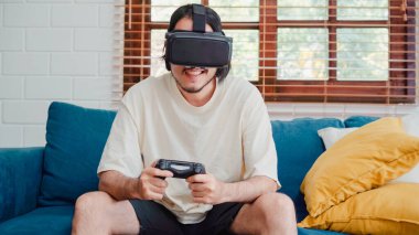 Asyalı bir adam sanal gerçeklik simülatörü ve joystick kullanarak oturma odasında video oyunu oynuyor, erkek evde kanepede uzanarak rahatlamak için kendini mutlu hissediyor. Erkekler ev kavramında oyun oynar rahatlar..