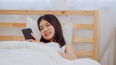Akıllı telefon kontrol sosyal medya kontrol kullanarak genç Asyalı kadın sabah uyandıktan sonra yatakta yatarken mutlu gülümseyerek duygu, Güzel çekici Japon kız evde yatak odasında dinlenmek gülümseyen.