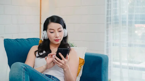 アジアの女性は音楽を聴いているし スマートフォンを使っている 自宅のリラックスした時間を使っている女性は 自宅のリビングルームでソファに横たわっている ヘッドフォンのコンセプトで幸せな女性のリスニング音楽 — ストック写真
