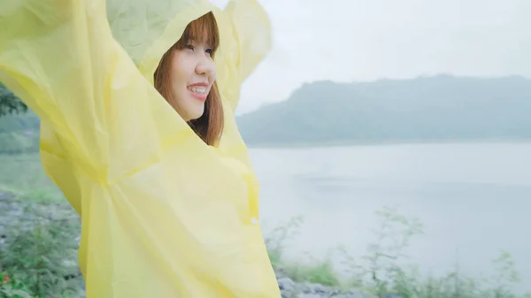 若いアジア女性感じ幸せな演奏の雨湖近く立っているレインコートを着用しながらの ライフ スタイルの女性と雨の日のリラックスをお楽しみいただけます — ストック写真