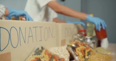 Bir grup genç Asyalı gönüllü, hayır kurumunda çalışırken yiyecek ve içecek paketleyip bağış kutularına bağışta bulunan eldiven giyiyor. Sosyal hizmet görevlisi, gıda bankası ve koronavirüs konsepti.