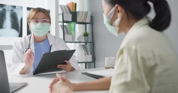 若いアジアの女性医師は ラップトップを使用して保護マスクを着用し 素晴らしいニューストークの結果や病院のオフィスでの少女患者との症状について議論を提供しています コロナウイルスの後のライフスタイルの新しい正常な — ストック動画