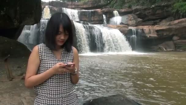 Mujer joven usando Smartphone al aire libre sentada cerca de la cascada y sonriendo — Vídeos de Stock