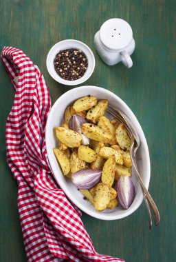 Oven-baked cut potato with red onion and Italian herbs, rustic, vintage or country style in a round bowl with red napkin on an old vintage green wooden background, top view
