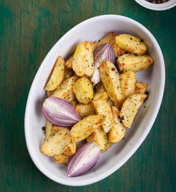 Oven-baked cut potato with red onion and Italian herbs, rustic, vintage or country style in a round bowl on an old vintage green wooden background, top view, with free space for text