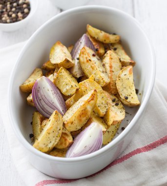 Oven-baked cut potato with red onion and Italian herbs, rustic, vintage or country style in a round bowl with white napkin on an old vintage wooden background, closeup