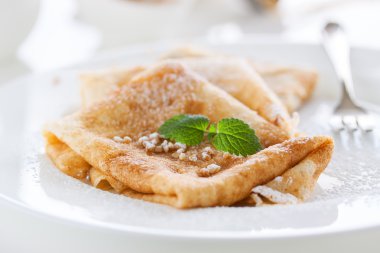 French crepes or pancakes with sugar powder, nuts and fresh mint on a white plate on a white background for breakfast, closeup