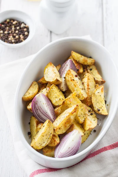 Oven-baked cut potato with red onion and Italian herbs, rustic, vintage or country style in a round bowl with white napkin on an kitchen background, top view