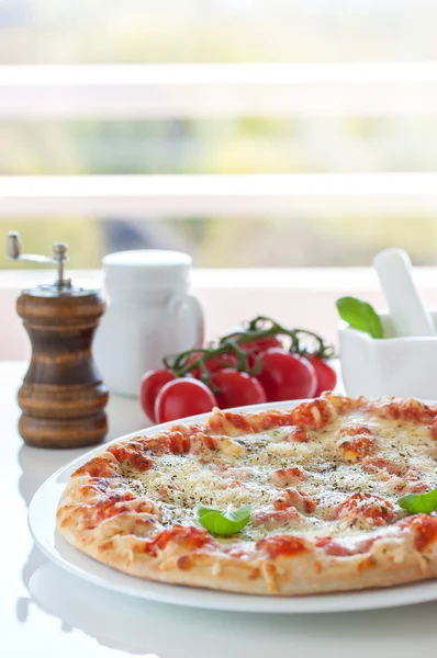 Свежая выпечка пиццы с расплавленным сыром, итальянские травы и томатный соус и свежий базилик на белой тарелке на кухонном фоне — стоковое фото