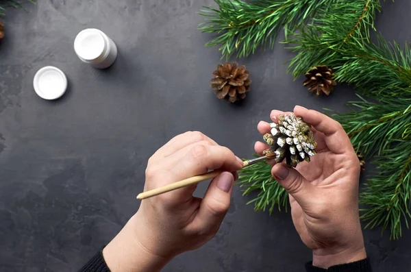Female hands painting pine cones for christmas decorations over dark background with pine tree branches.