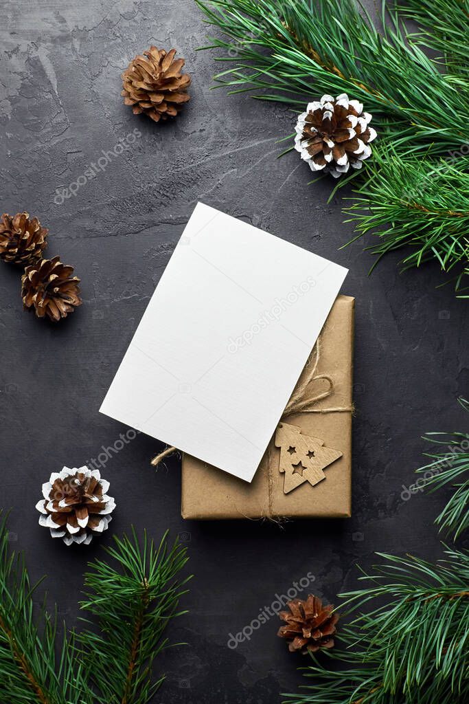 Greeting card mockup with christmas gift box and pine tree branches with cones on dark stony background. Winter holidays flat lay, top view, copy space.