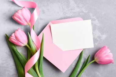 Pembe zarflı tebrik kartı modeli ve gri beton arka planda kurdeleli lale çiçekleri, üst görünüm, kopyalama alanı