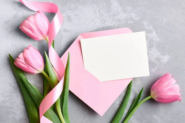 Pembe zarflı tebrik kartı modeli ve gri beton arka planda kurdeleli lale çiçekleri, üst görünüm, kopyalama alanı