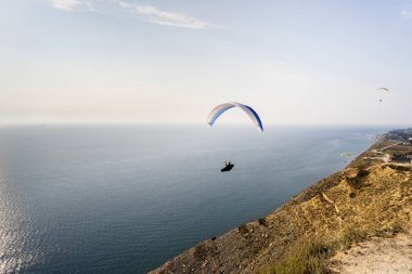 Güneşli bir yaz gününde mavi gökyüzünde dağların ve denizin üzerinden bir paraglider uçar. Parlak mavi deniz ve yeşil çimen, yaz, özgürlük, macera, eğlence.