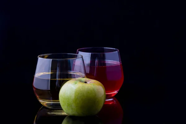 Two glasses of juice and an apple on a black background.