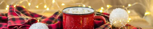 Banner of Christmas coffee in a red cup with marshmallows and checked plaid. New year concept decorated white balls candy cane and light garland bokeh