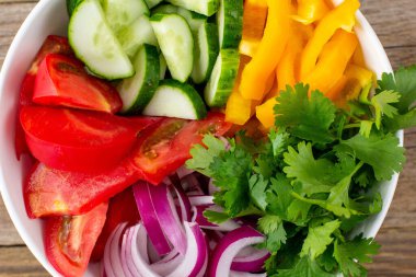 Farklı sebzelerle dolu bir tabak gökkuşağı salatası ve ahşap arka planda beyaz kasede otlar. Üst görünüm