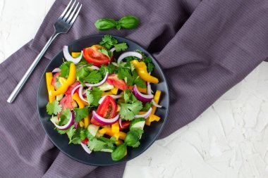 Farklı sebzelerle dolu bir tabak gökkuşağı salatası ve beyaz taş zemin üzerinde çatal ve peçeteli siyah tabakta otlar. Kopyalama alanı olan üst görünüm