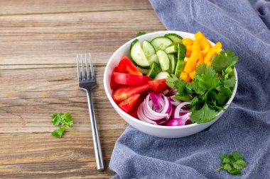 Farklı sebzelerle dolu bir tabak gökkuşağı salatası ve doğal ahşap zemin üzerine çatal ve peçete ile beyaz kâse içinde otlar.