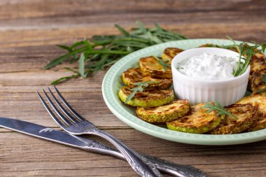 Çatal ve bıçakla tabakta ekşi krema soslu kızarmış kabak dilimleri. Doğal kahverengi ahşap arka planda akşam yemeği için otlu kızarmış kabaklar..