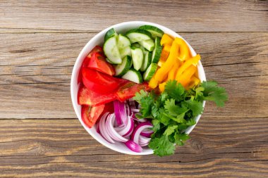 Farklı sebzelerle dolu bir tabak gökkuşağı salatası ve ahşap arka planda beyaz kasede otlar. Üst görünüm