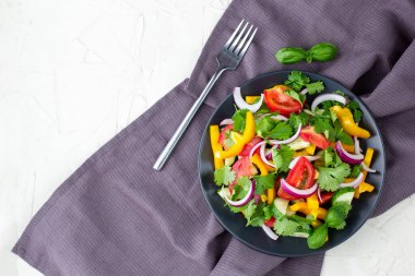 Farklı sebzelerle dolu bir tabak gökkuşağı salatası ve beyaz taş zemin üzerinde çatal ve peçeteli siyah tabakta otlar. Kopyalama alanı olan üst görünüm