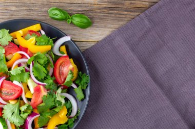 Farklı sebzelerle dolu bir tabak gökkuşağı salatası ve doğal ahşap arka planda peçeteli siyah tabakta otlar. Kopyalama alanı olan üst görünüm