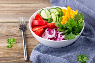 Farklı sebzelerle dolu bir tabak gökkuşağı salatası ve doğal ahşap zemin üzerine çatal ve peçete ile beyaz kâse içinde otlar.