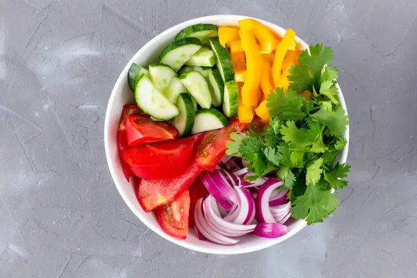 Farklı sebzelerle dolu bir tabak gökkuşağı salatası ve gri taş arka planda beyaz kâse içinde otlar. Üst görünüm