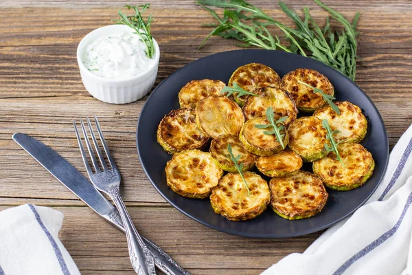 Çatal ve bıçakla tabakta ekşi krema soslu kızarmış kabak dilimleri. Doğal kahverengi ahşap arka planda akşam yemeği için otlu kızarmış kabaklar..