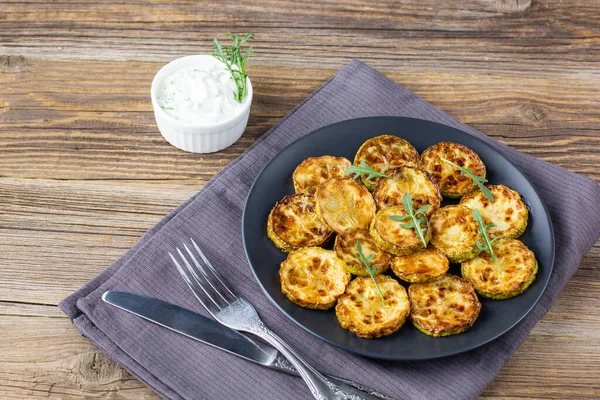 Çatal ve bıçakla tabakta ekşi krema soslu kızarmış kabak dilimleri. Doğal kahverengi ahşap arka planda akşam yemeği için otlu kızarmış kabaklar..