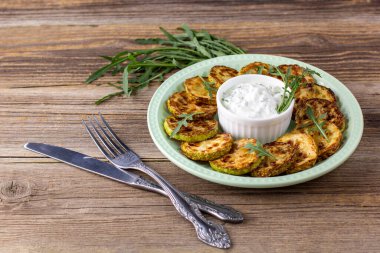 Çatal ve bıçakla tabakta ekşi krema soslu kızarmış kabak dilimleri. Doğal kahverengi ahşap arka planda akşam yemeği için otlu kızarmış kabaklar..
