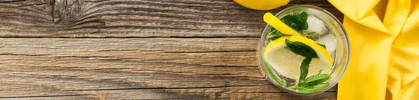 banner of Lemonade or mojito cocktail with lemon and mint with ice. Cold summer refreshing drink decorated with yellow napkin on wooden background. Top view with copy space