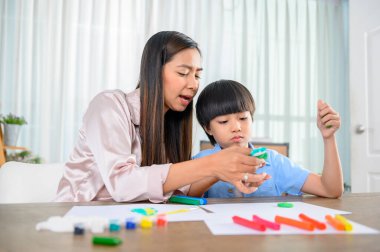 Asyalı anne oğluyla birlikte evde çalışıyor. Anne ve çocuk hamur oynuyorlar. Çocuk plasticine kil modeli yaratıyor. Kadın yaşam tarzı ve aile aktiviteleri.