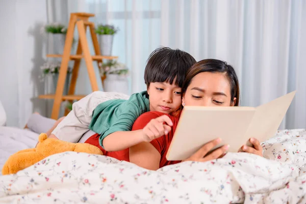 Asian mother work home together with son. Mom and kid reading fairy tale before sleep on the bed. Woman lifestyle and family activity.