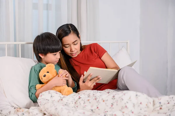 Asian mother work home together with son. Mom and kid reading fairy tale before sleep on the bed. Woman lifestyle and family activity.