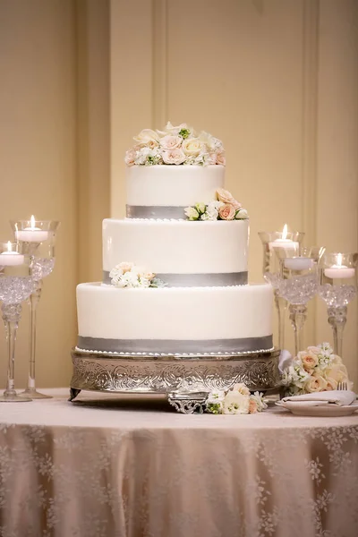 Wedding ceremony three tiered cake made with white frosting and grey trim decorated with white and pink roses on a table covered with silk tablecloth